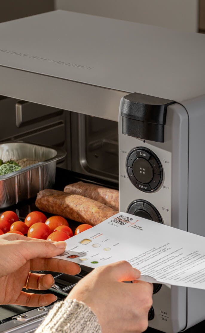 Tovala Smart Oven with broccoli, tomatoes, and sausage inside and a person scanning the meal card QR code to cook the meal.